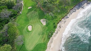 Una vista aerea di un campo da golf accanto a una spiaggia con dune sabbiose e onde dell'oceano.