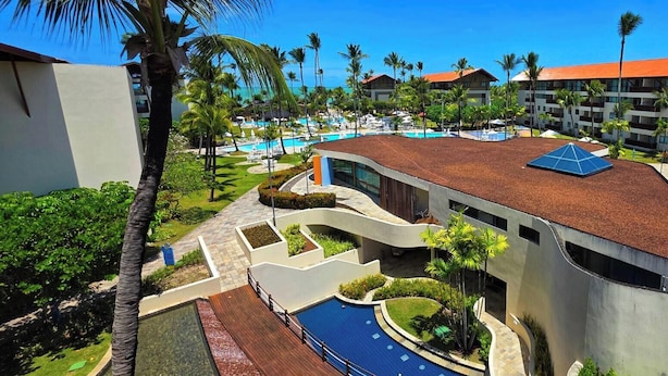 A resort with a swimming pool, palm trees, and a modern building with a curved roof. - Marulhos Suítes Resort (Ipojuca)