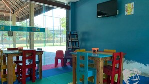 A room with a blue wall, wooden tables and chairs, a television, and a window with a view of an outdoor area. - Marulhos Suítes Resort (Ipojuca)