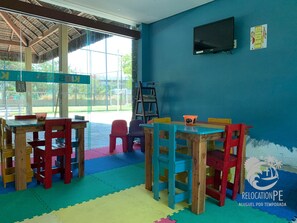 A room with a blue wall, wooden tables and chairs, a television, and a window with a view of an outdoor area. - Marulhos Suítes Resort (Ipojuca)