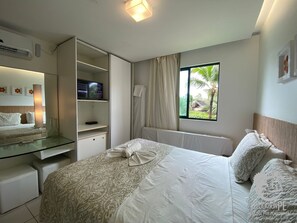 A hotel room with a bed, a TV, a wardrobe, a window with a view of palm trees, and a glass table. - Marulhos Suítes Resort (Ipojuca)