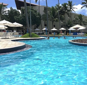 A swimming pool with lounge chairs and umbrellas, surrounded by palm trees and a building. - Marulhos Suítes Resort (Ipojuca)
