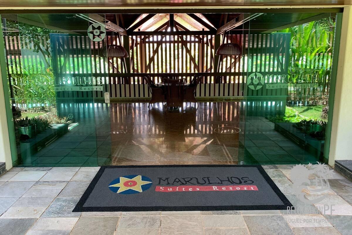 Entrada de hotel con puerta de cristal, felpudo con el nombre del hotel y cerca de madera con plantas.