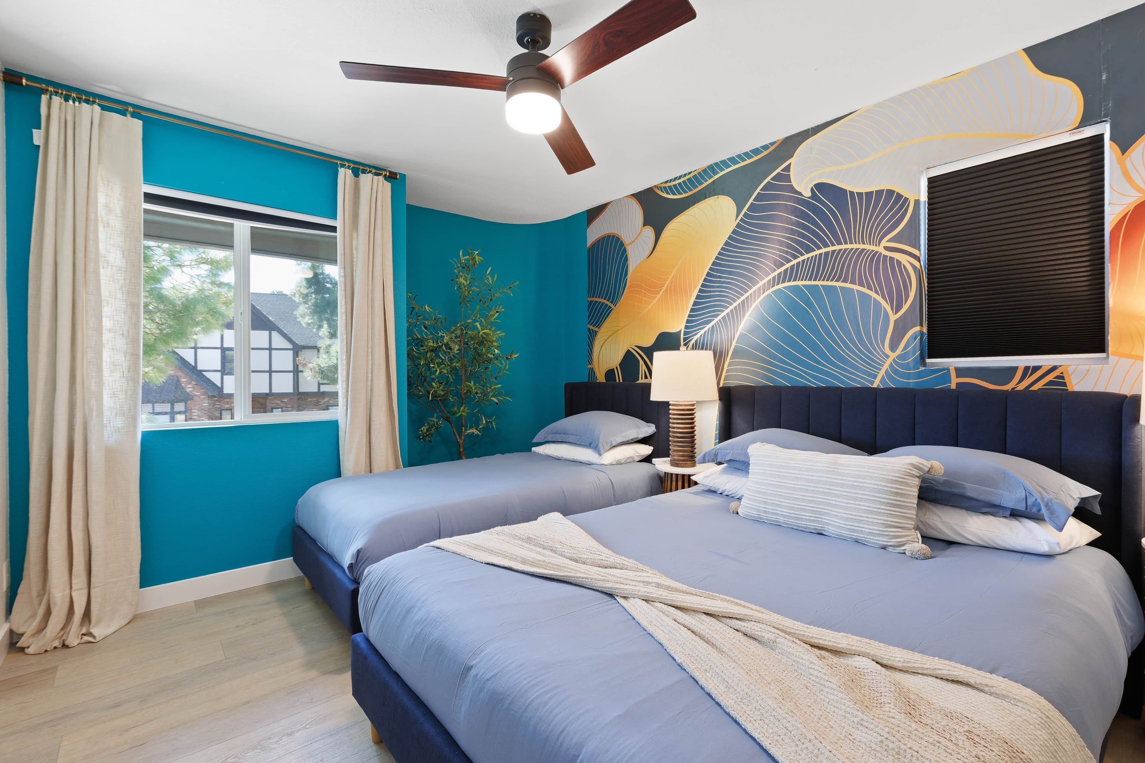 A bedroom with two beds, a ceiling fan, a window with curtains, a wall mural, and a bedside table with a lamp.