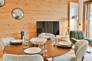 Ein Essbereich mit einem Holztisch, gedeckt für vier Personen, umgeben von gepolsterten Stühlen. Ein Fernseher ist an der Wand montiert, und im Hintergrund ist eine Lampe zu sehen.