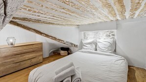 Une chambre avec un plafond en pente, une commode en bois, un lit avec du linge de lit blanc et une étagère fixée au mur.
