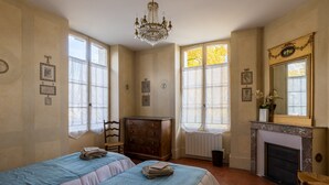 Une chambre avec deux lits, une commode, un lustre, une cheminée et de grandes fenêtres avec des rideaux.