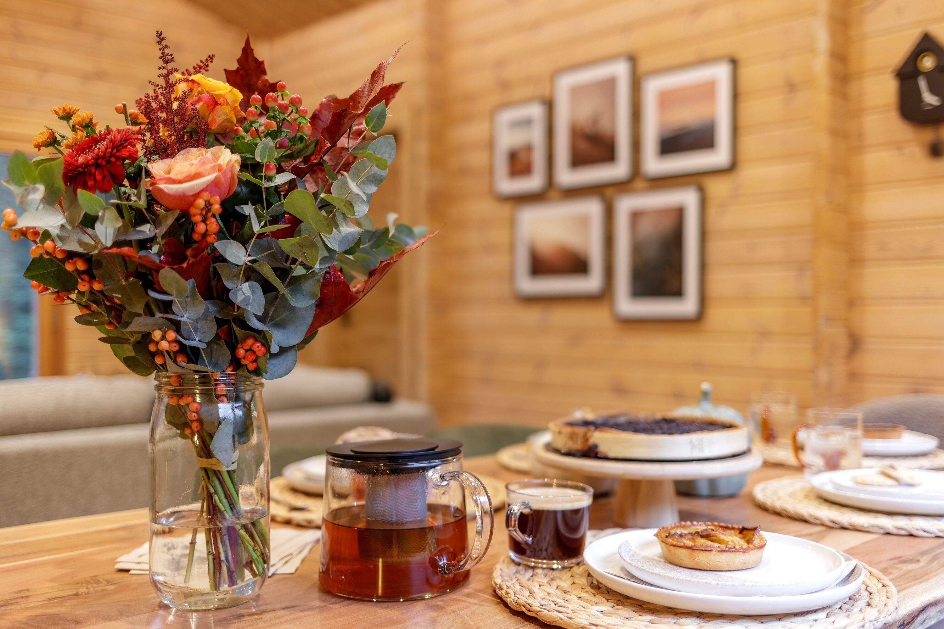 Un intérieur en bois avec une table dressée pour le thé et les pâtisseries, un vase rempli de fleurs et des tableaux encadrés accrochés au mur.
