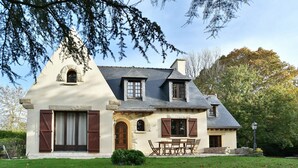 Une maison à deux étages, dotée d’une façade en pierre, d’un toit en pente et d’un petit porche à l’avant, avec une table et des chaises.