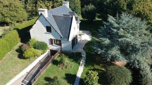 Une grande maison blanche à deux étages, dotée d’un toit à forte pente, entourée d’une végétation luxuriante et d’arbres.