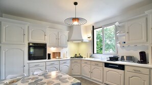 Une cuisine avec des meubles blancs, une table à manger recouverte d’une nappe à motifs de feuilles, un four encastré et une fenêtre donnant sur un paysage verdoyant.