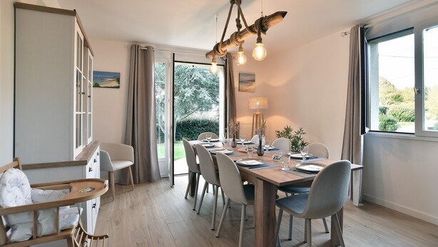 Un coin repas avec une table en bois, dressée pour quatre personnes, entourée de chaises. Une grande fenêtre offre une vue sur la verdure environnante.