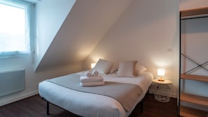 Een slaapkamer met een schuine kap, een bed met witte beddengoed, twee kussens, een nachtkastje met een lamp en een radiator.