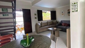 A living area with a sofa, a dining table with chairs, a staircase, and a view of the outside. - Condomínio Merepe Residence (Ipojuca)