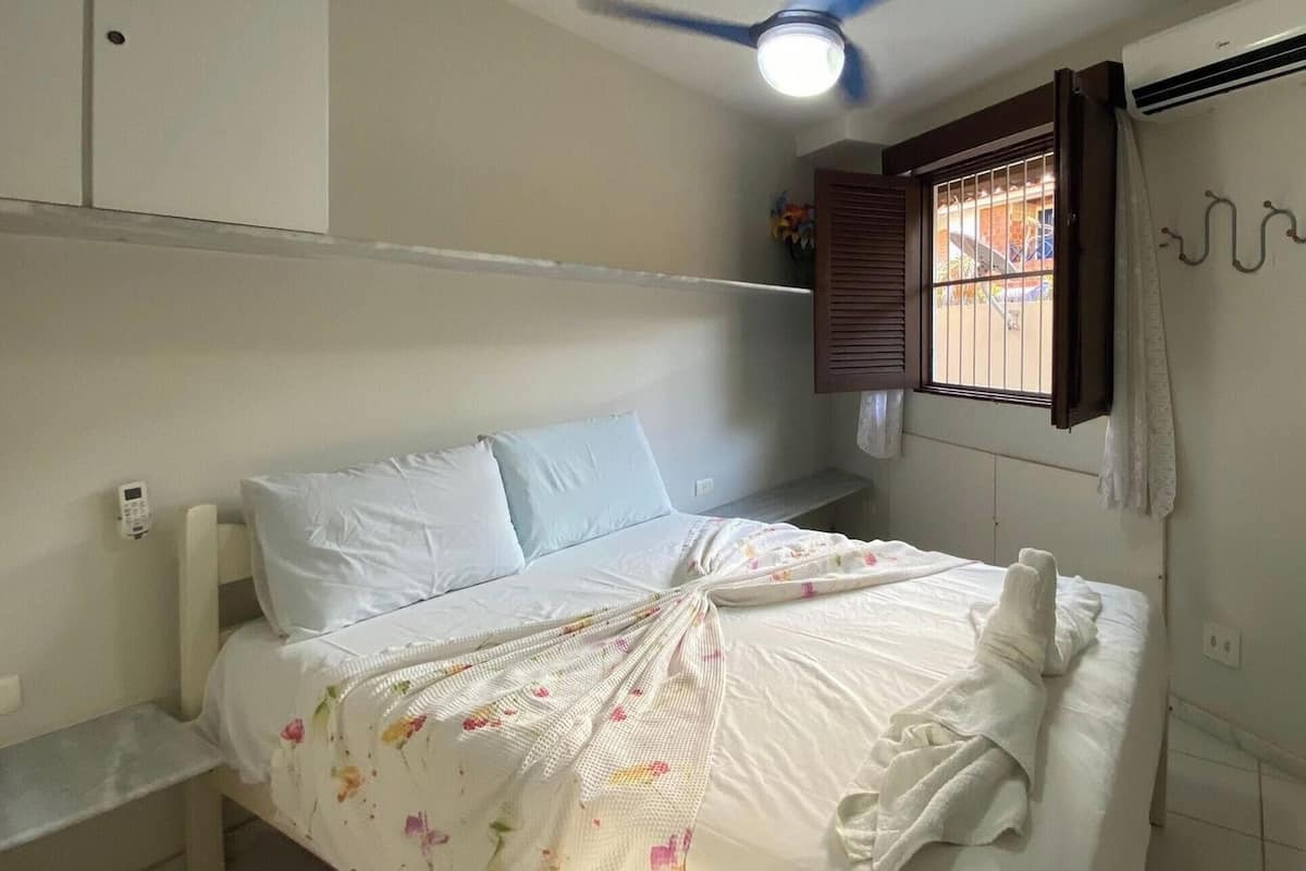 Un dormitori amb un llit, un ventilador de sostre, una finestra amb reixes i un aire condicionat fixat a la paret.