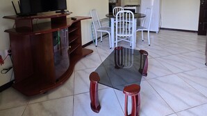 Ein Wohnzimmer mit Fernseher auf einem Holzboden, einem Esstisch mit weißen Stühlen und einer Küchenzeile mit Kühlschrank und Mikrowelle.