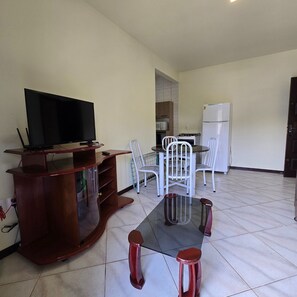 A living room with a TV on a wooden stand, a dining table with white chairs, and a kitchen area with a fridge and microwave. - Residencial 01 – Your perfect accommodation close to the beach. (Bombinhas)