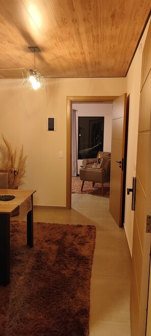 A hallway with a wooden ceiling, a dining area with a table and chairs, and a view into a living room with a sofa and a bottle. - Romantic apartment with hot tub and mountain views, breakfast included. (Campos do Jordão)