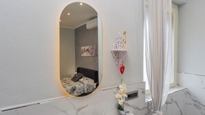 A bathroom with a marble floor, a round mirror, a bed with a floral bedspread, a wall-mounted shelf with decorative items, and a window with curtains. - Fabiola Guest House (Pompei)