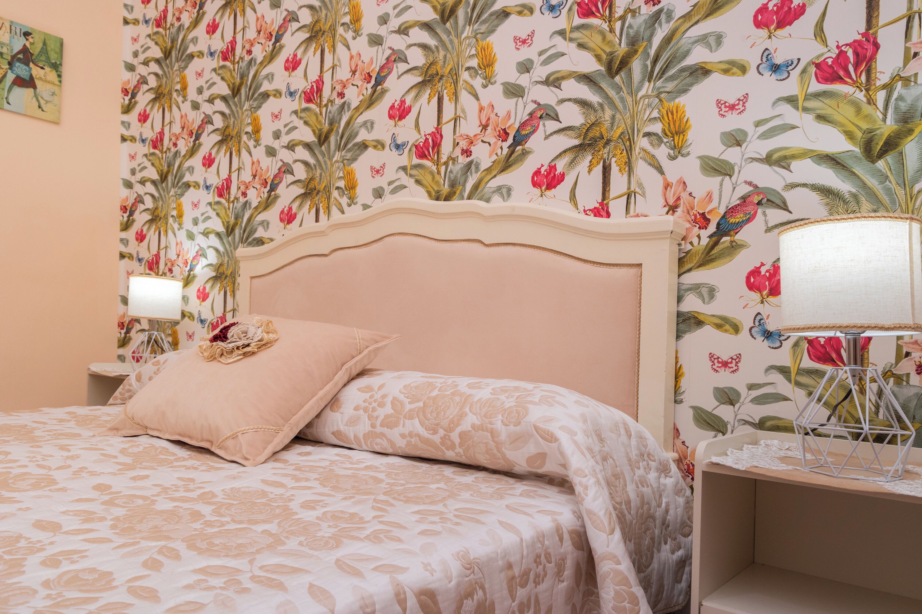 Un dormitorio con papel tapiz floral, una cama con cubrecama floral en tonos beige y blanco, una mesita de noche con lámpara y un estante en la pared con un objeto decorativo.