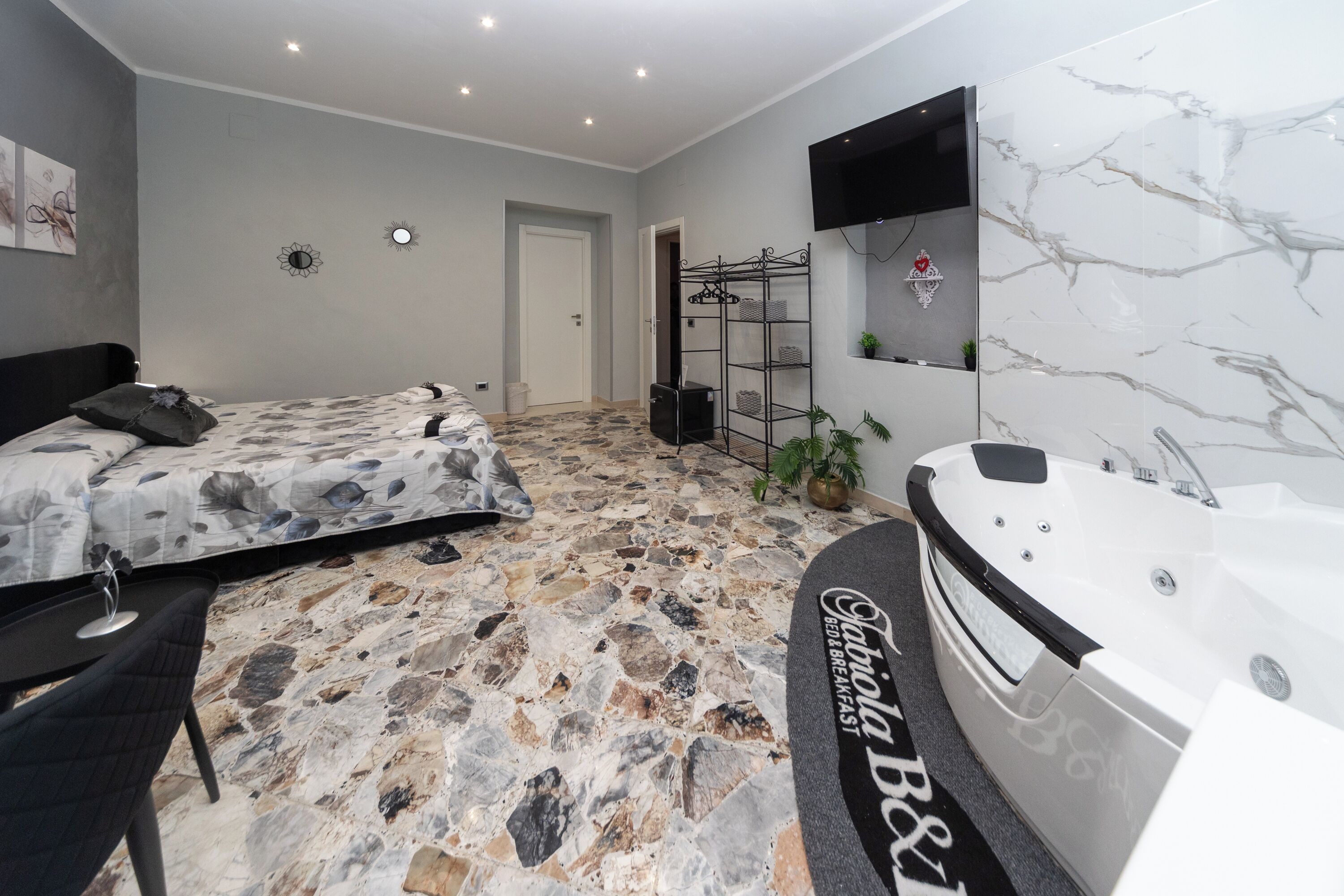 A bedroom with a stone-patterned floor, a bed with a floral duvet, a black chair, a black speaker, a white bathtub, and a wall-mounted TV.