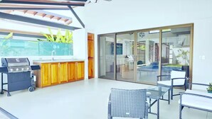 Un área de cocina exterior moderna con una parrilla, armarios de madera y un ventanal que da a una piscina.