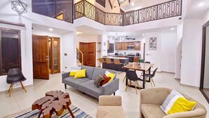 Un salón amplio con un sofá gris, sillones color beige y una mesa de comedor. Una escalera conecta el salón con una cocina y zona de comedor.