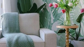 Ein Wohnzimmer mit einem beigefarbenen Sofa, einer grünen Strickdecke, einem Holznachttisch mit Blumen und einer großen Pflanze im Hintergrund.