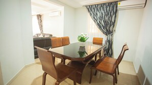 Een eetgedeelte met een glazen tafel en houten stoelen, een kamerplant en uitzicht door een raam met gordijnen.