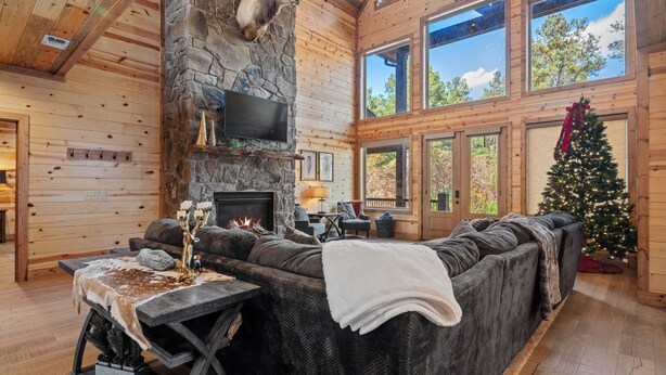 A cozy log cabin interior with a stone fireplace, a large sectional sofa, a flat-screen TV mounted on the wall, and a Christmas tree. - Spacious 5-bedroom cabin Centrally located in Hochatown with Backup Generator (Broken Bow)