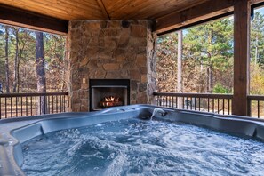 Un bain à remous avec une cheminée en pierre, entouré d’une terrasse en bois et offrant une vue sur les arbres.
