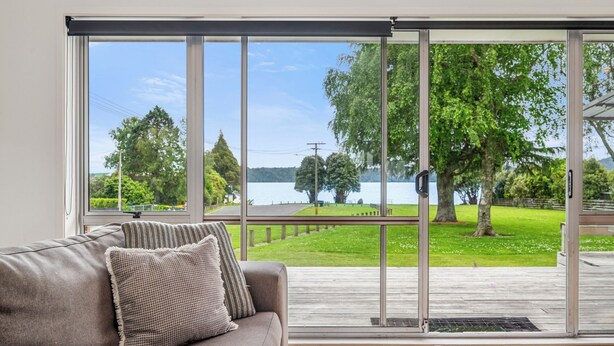 A room with a large sliding glass door leading to a deck with a view of a lake and trees. - Rotoiti Escape - Lake Rotoiti Holiday Home (RD 4)