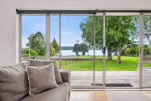 A room with a large sliding glass door leading to a deck with a view of a lake and trees. - Rotoiti Escape - Lake Rotoiti Holiday Home (RD 4)