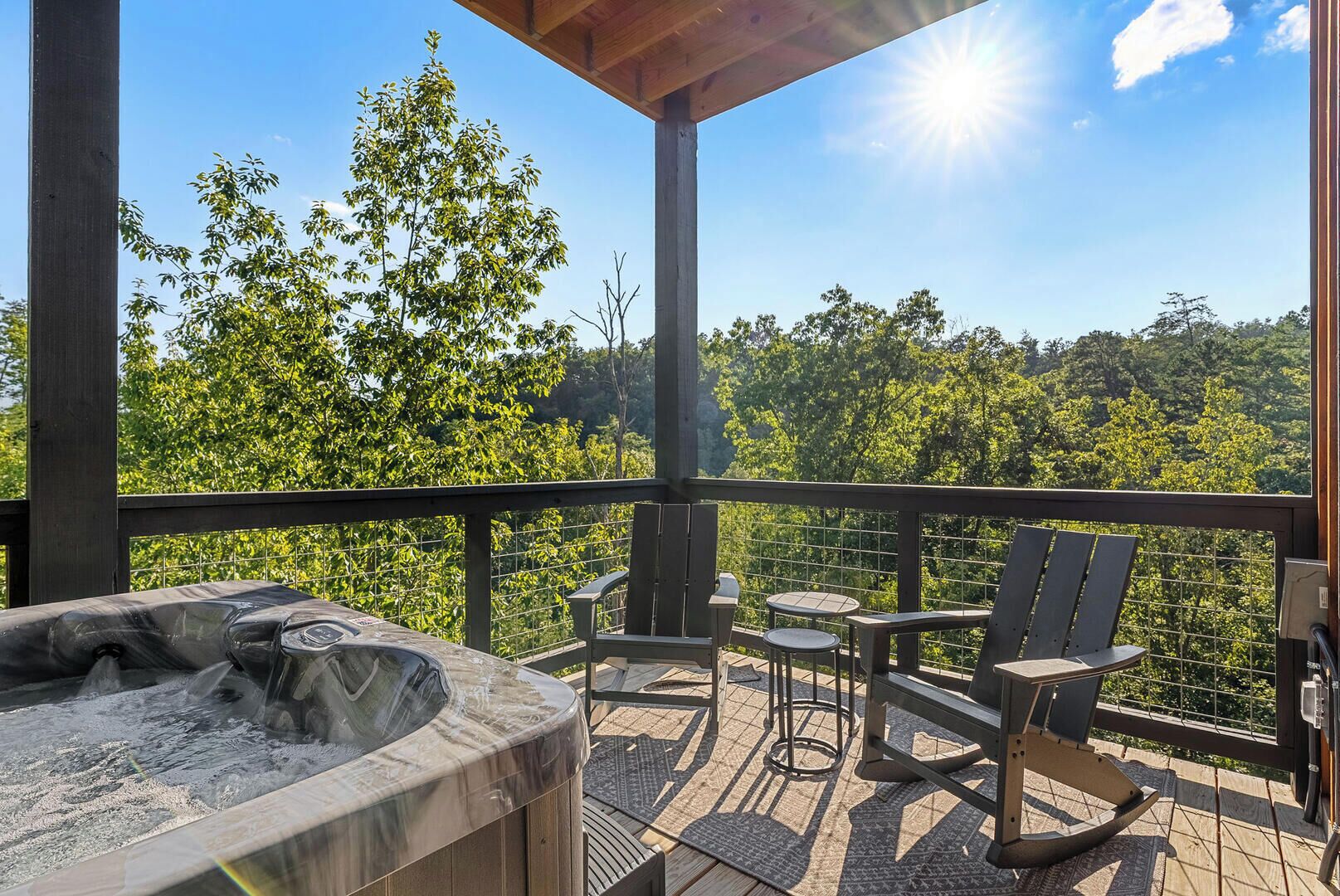 Un bain à remous sur une terrasse en bois, agrémentée de chaises et de tables, entourée d’une rambarde en métal, et offrant une vue sur une zone boisée.
