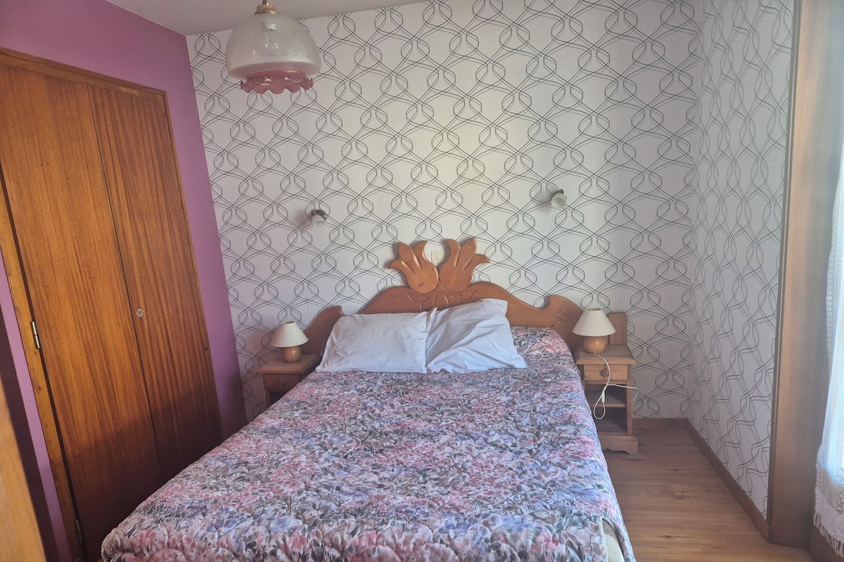 Quarto com cama de madeira, colcha florida, duas luminárias de cabeceira, guarda-roupa de madeira e papel de parede estampado.