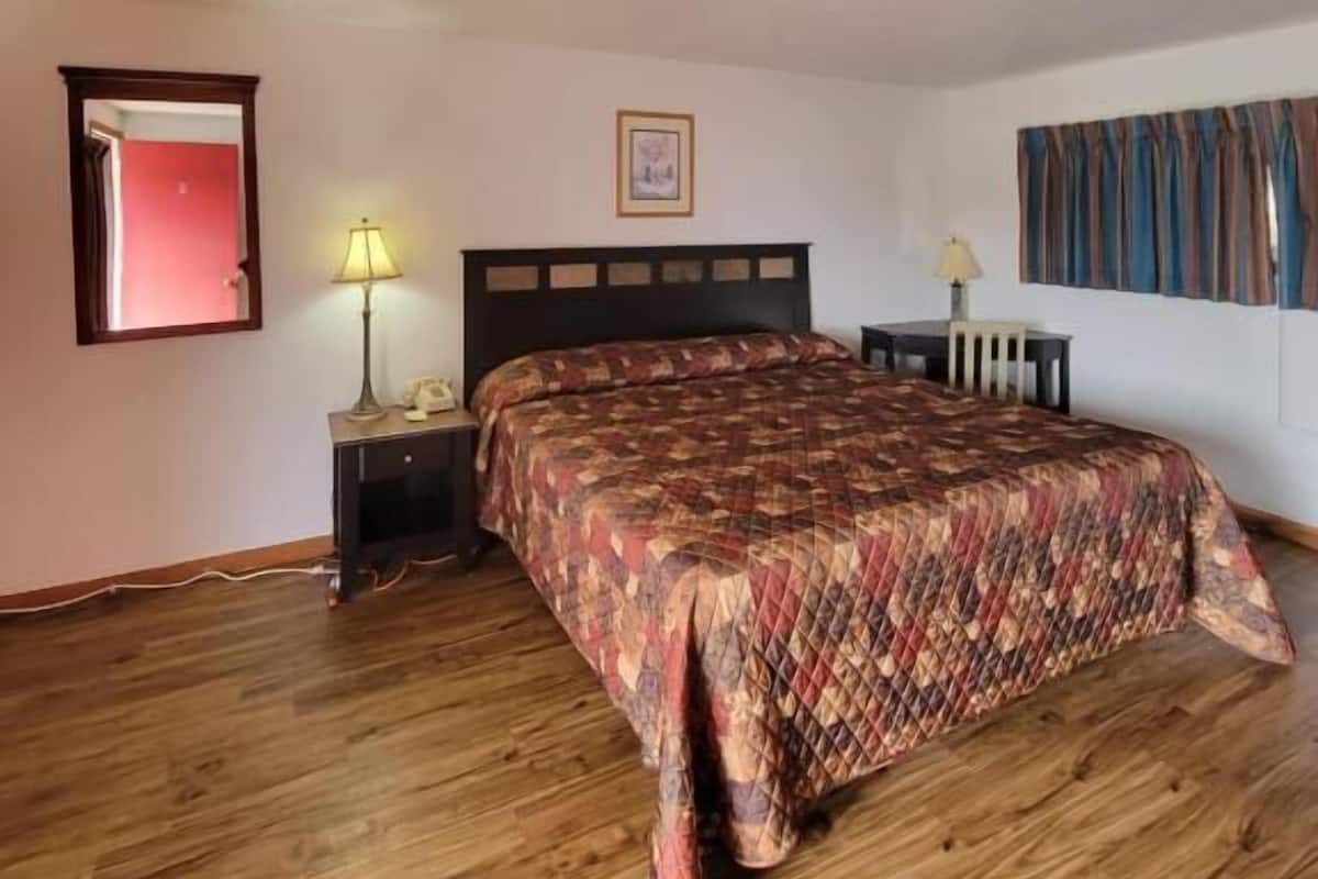A bedroom with a wooden floor, a bed with a patterned bedspread, a nightstand with a lamp, a framed picture on the wall, and a window with curtains.