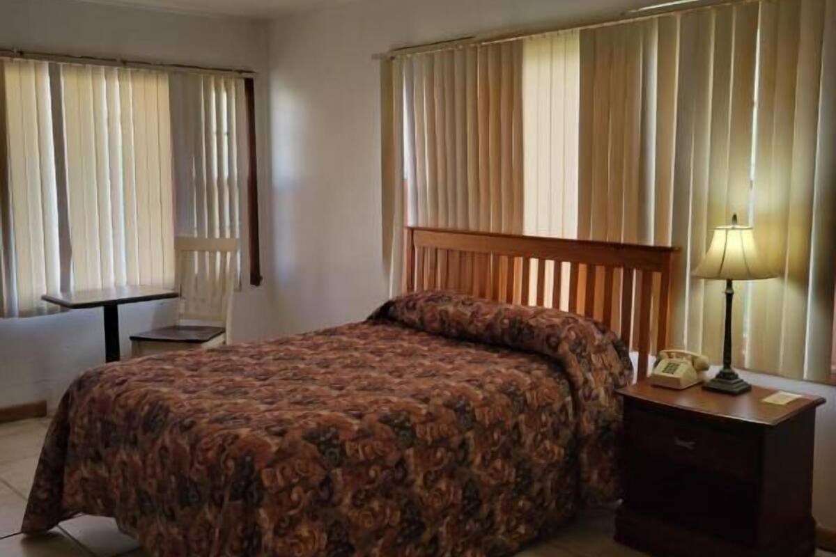 A hotel room with a bed, a nightstand with a lamp, a chair, and a window with curtains.
