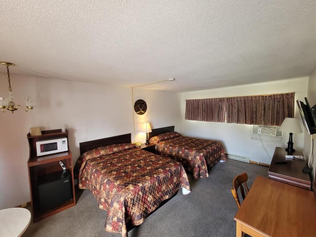 A hotel room with two beds, a desk, a chair, a microwave, a lamp, and a clock on the wall.