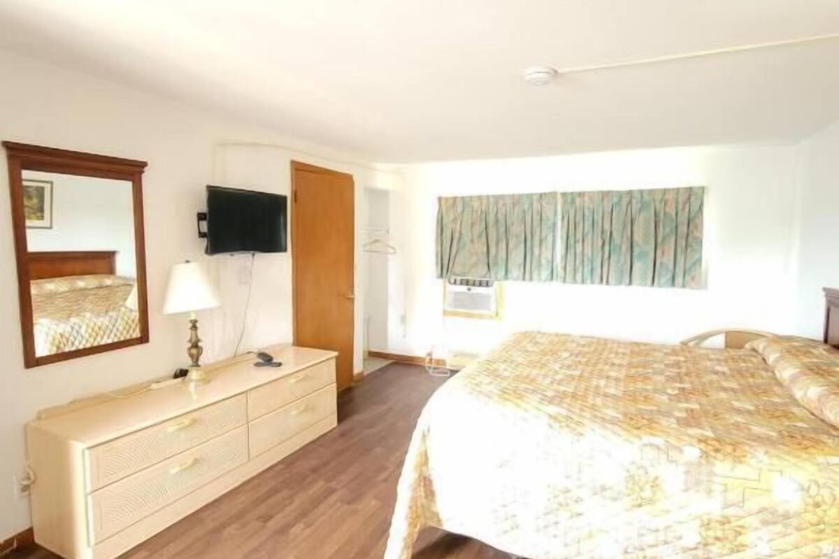 A bedroom with a wooden floor, a bed with a patterned bedspread, a dresser, a mirror, a television, and a window with curtains.