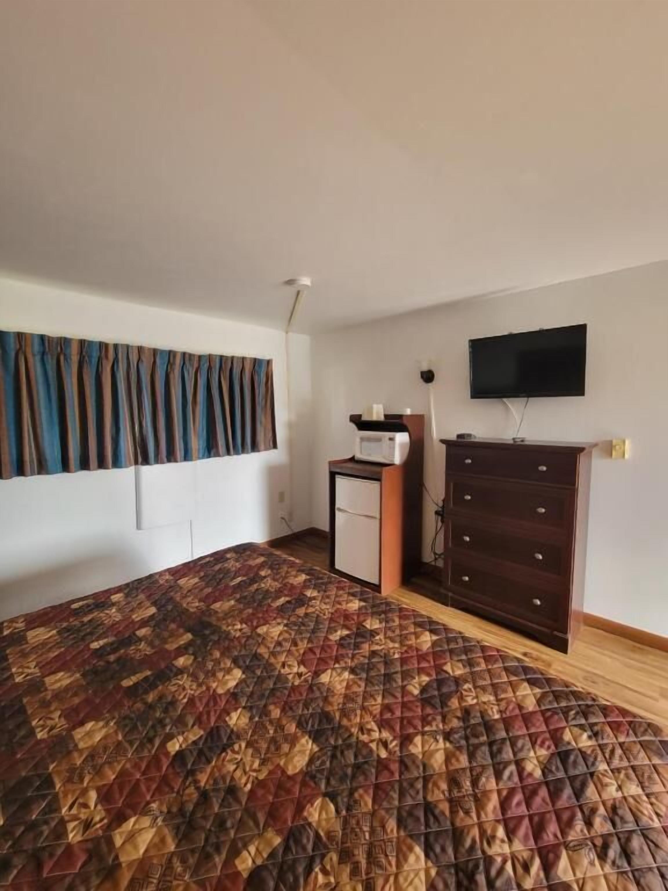 A room with a patterned bedspread, a small refrigerator, a TV mounted on the wall, and a wooden dresser.