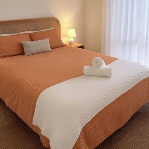 Una cama bien hecha con una colcha blanca y una toalla enrollada, una lámpara de noche y una ventana con cortinas.