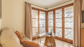 Uma sala de estar com sofá, uma mesa e um laptop, e janelas amplas com cortinas.