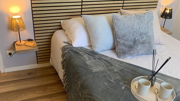 Un lit bien fait, avec une couverture grise, deux oreillers et un plateau sur lequel se trouvent deux tasses et une cuillère.
