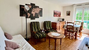 A living room with a sofa, two striped armchairs, a wooden dining table with chairs, a small round table with a doily, a world map on the wall, a ceiling fan, and a window with a view of greenery. - Bright T2 with Wi-Fi and washing machine, center of La Roche-Posay (La Roche-Posay)
