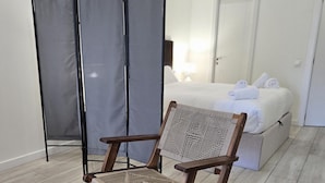 Ein hölzerner Stuhl mit gewebter Sitzfläche und Lehne, ein Bett mit weißen Leinen und Handtüchern sowie ein grauer Vorhang in einem Hotelzimmer.