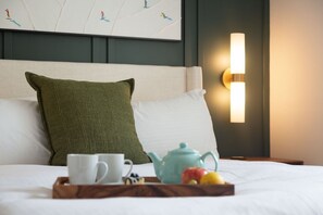 Un lit bien fait, avec un oreiller blanc et un oreiller vert, un plateau avec une théière, une tasse et des fruits, et un luminaire fixé au mur.