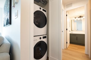 Une buanderie équipée de deux machines à laver à chargement frontal, un couloir menant à une salle de bain avec un meuble-lavabo et un miroir, et un canapé de couleur claire au premier plan.