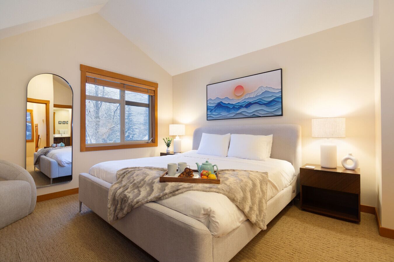 A bedroom with a large bed, a window with a view of trees, a painting of mountains, and a bedside table with a lamp.