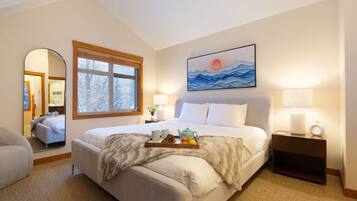 A bedroom with a large bed, a window with a view of trees, a painting of mountains, and a bedside table with a lamp.