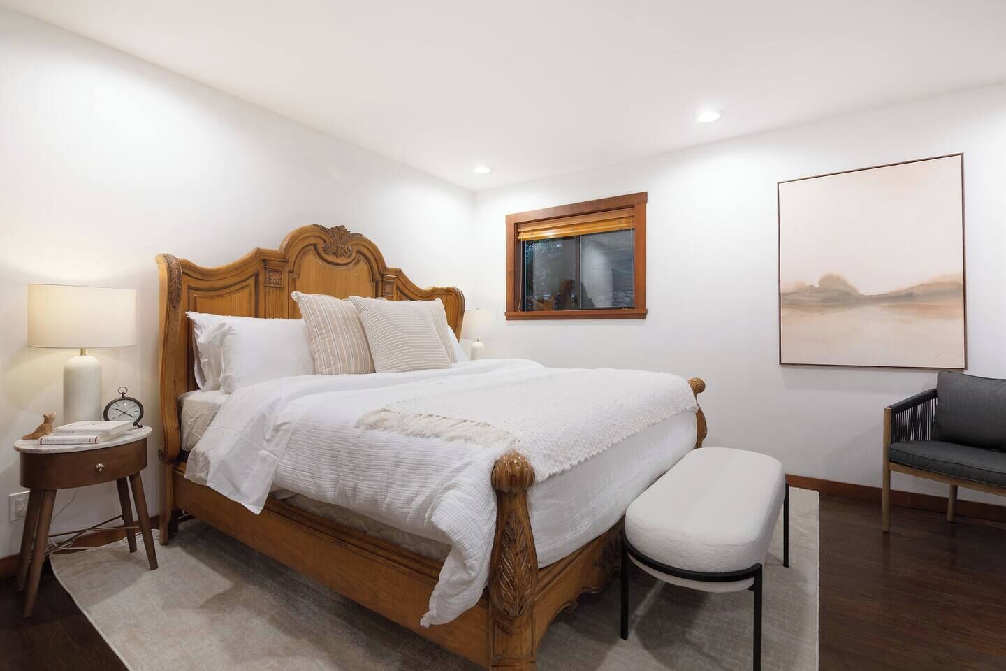 A neatly arranged bedroom with a wooden bed, a bedside table with a lamp, a chair, and a framed picture on the wall.